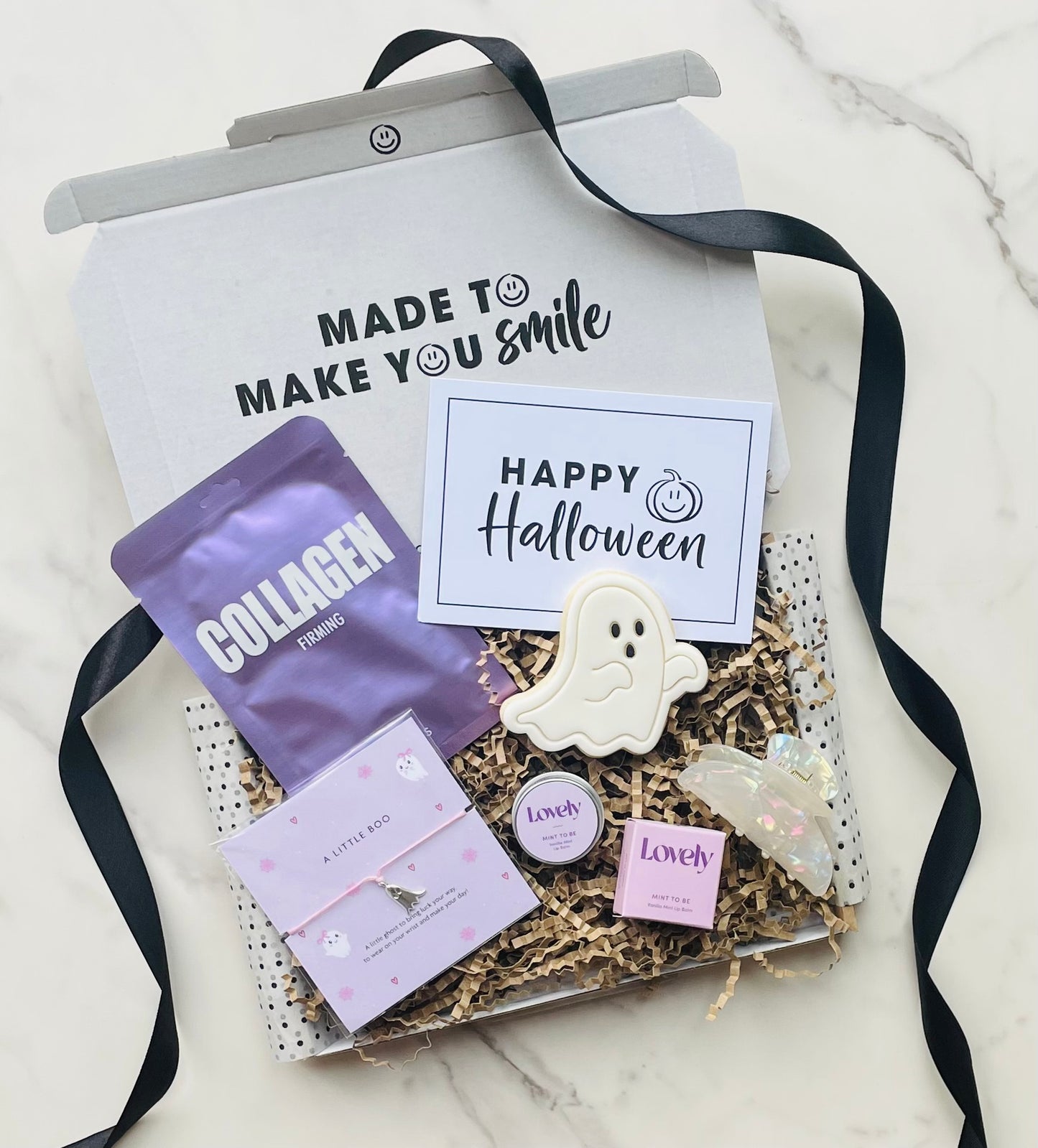 Letterbox gift with Halloween-themed items including a collagen pack and ghost-shaped cookie on a white marble surface.