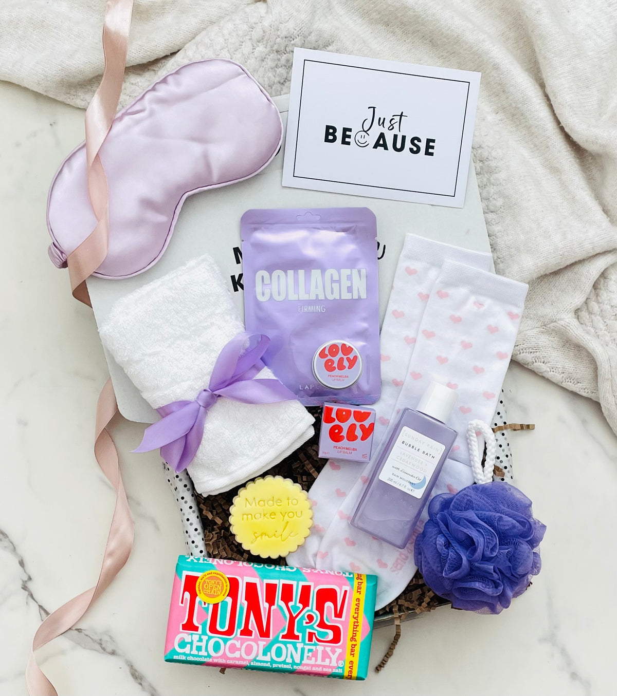 A curated gift basket arrangement on a marble surface with a cream blanket. The set includes a lavender satin eye mask with a pink ribbon, white hand towels tied with a purple ribbon, a purple collagen pouch, pink heart-print socks, a small lavender bottle, a purple loofah, a yellow sponge, a Tony's chocolate bar, and a card that reads 'Just BECAUSE'.