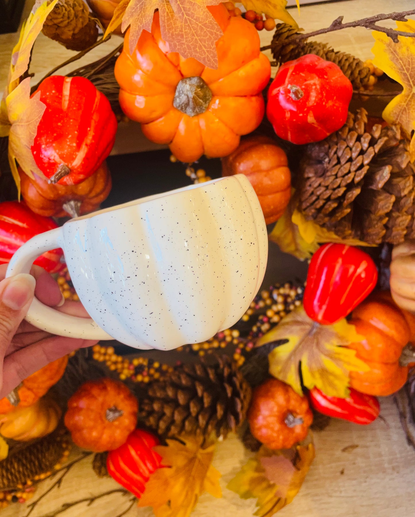 A person holds n adorable Halloween pumpkin mug in front of a decorative wreath, showcasing festive autumn spirit.