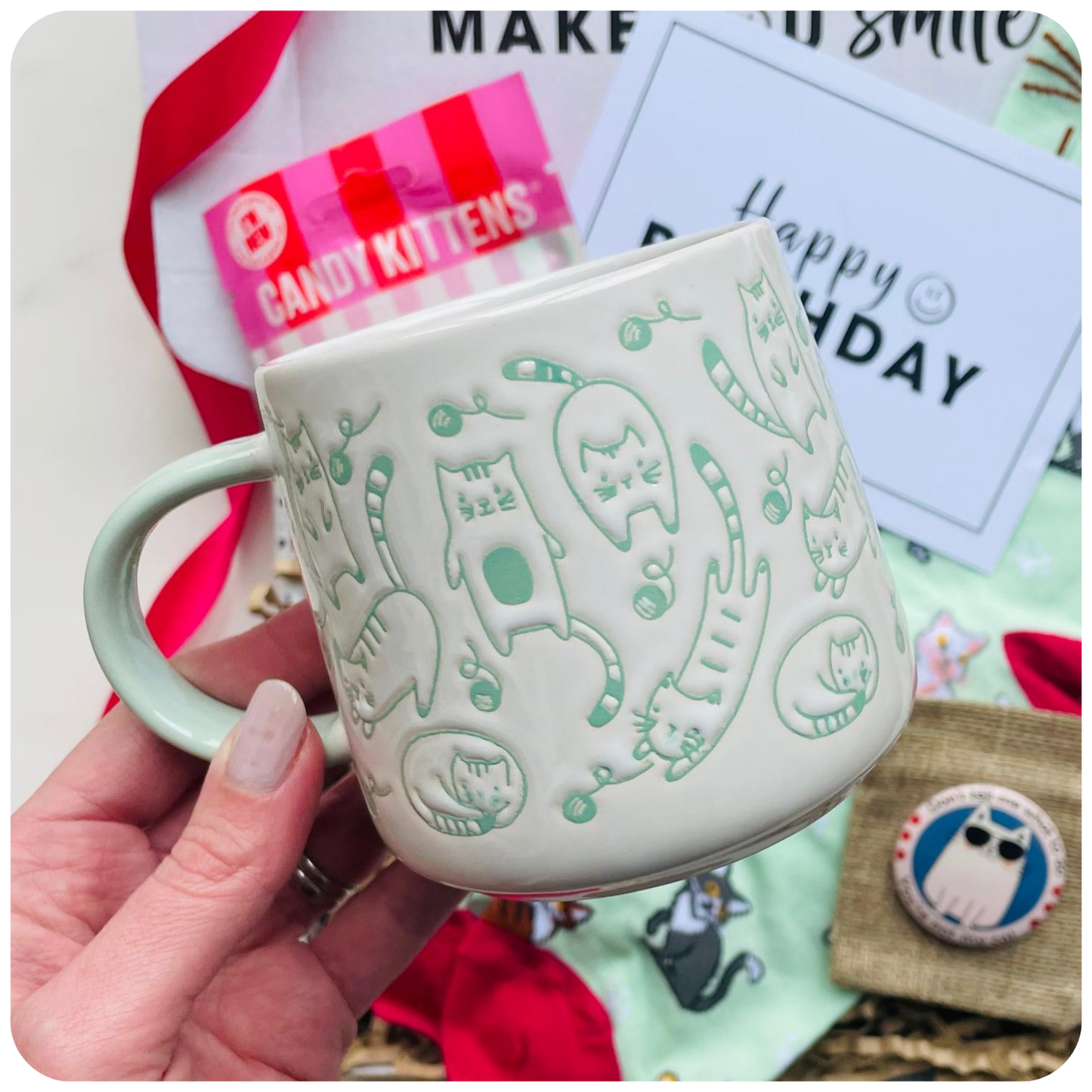 White mug with cat design held by a hand, surrounded by gift cards and candy. Cat theme gift